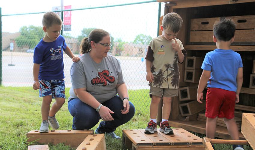 Three little boys and a teacher play with large hallow wooden outdoor blocks.